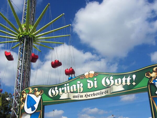 Eingangsschild zum Herbstfest mit der Begrüßung "Grüß Gott" und einem hohen Kettenflieger im Hintergrund