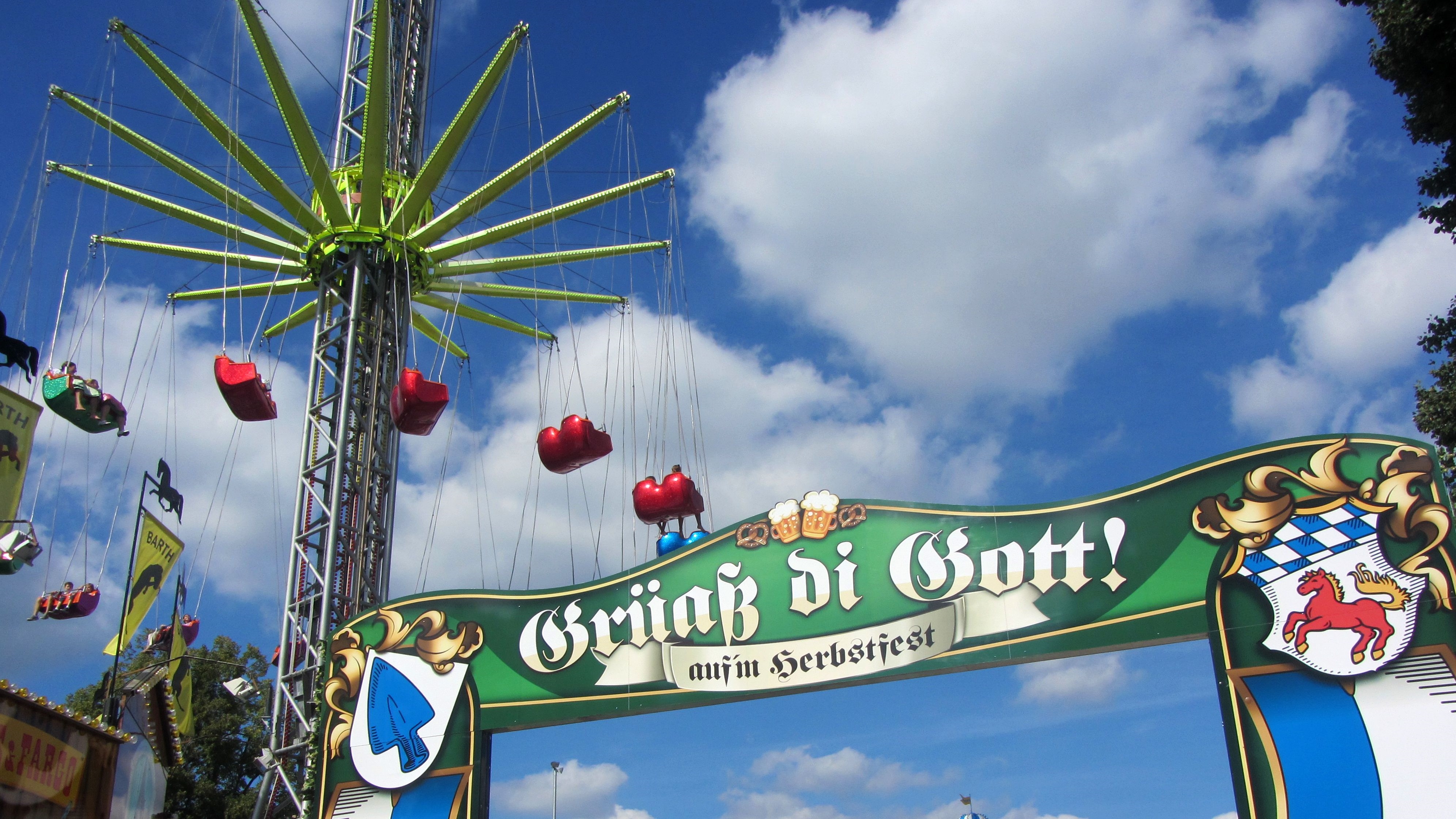 Eingangsschild zum Herbstfest mit der Begrüßung "Grüß Gott" und einem hohen Kettenflieger im Hintergrund